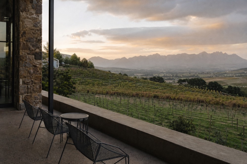 A stone-and-glass tasting terrace looking out over terraced vineyard rows toward a distant valley at golden hour, empty chairs and a low wall, no branding, no text, no people
