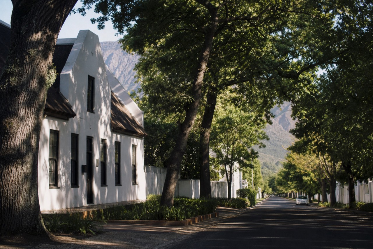 A quiet oak-lined street in a small historic town with whitewashed gabled buildings and a mountain rising at the far end, dappled shade on the pavement, no signage, no text, no people