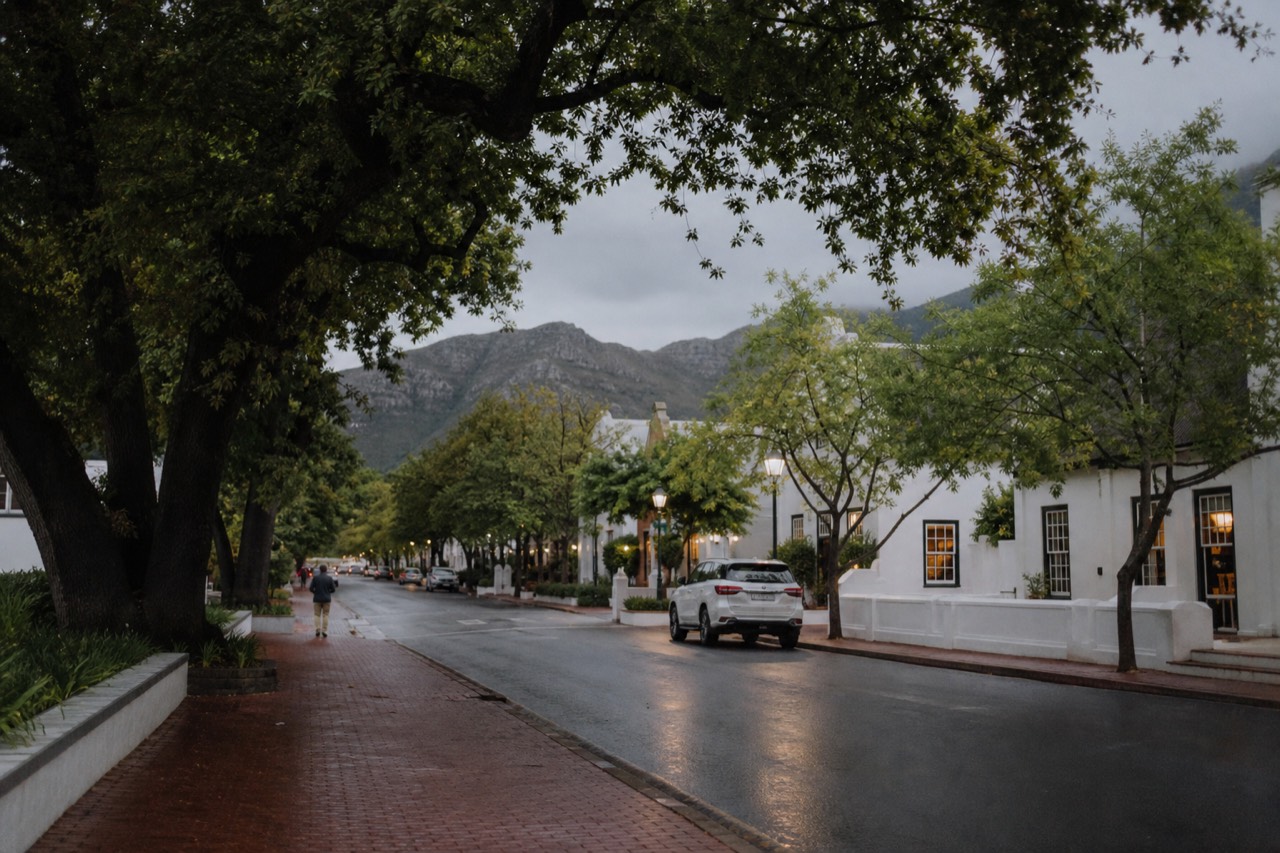 An oak-lined winelands town street of whitewashed Cape Dutch buildings at dusk with warm window light and mountains behind, no readable signs, no text, no faces