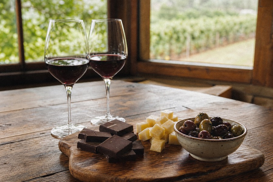 A wine-pairing board of local chocolate, cheese and olives beside two glasses of red on a rustic table, vineyard light coming through