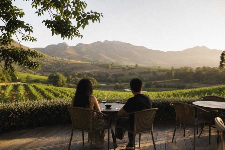 A couple at an unhurried private tasting on a quiet winery terrace, glasses of red wine on the table, oak-lined vineyard and mountains behind