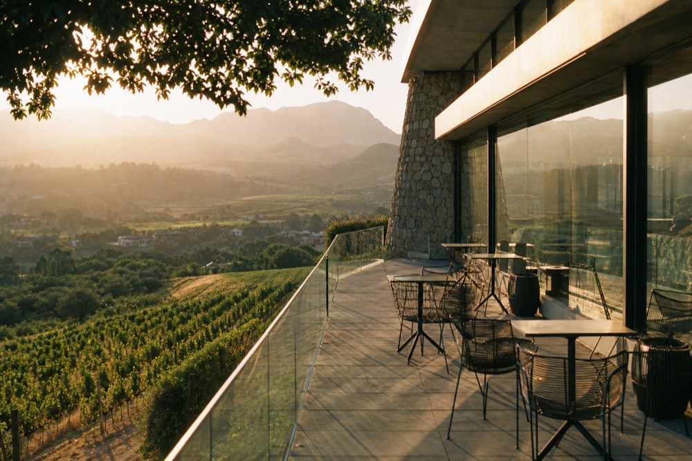 A modern glass-and-stone tasting terrace overlooking terraced vineyards and a distant valley at golden hour, empty chairs, no branding, no text, no people