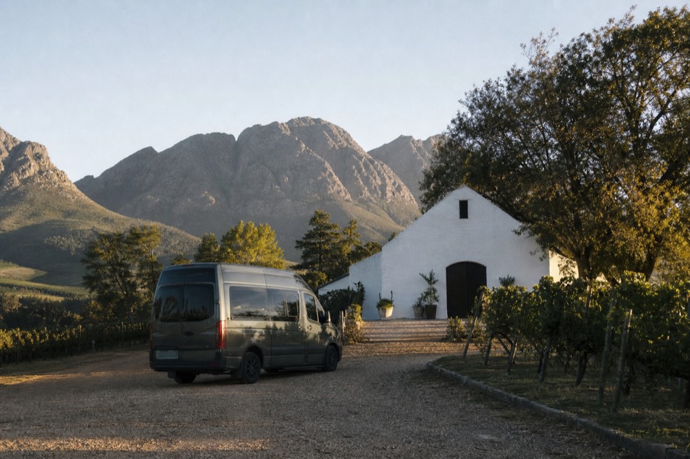 A small touring van parked at the end of a gravel drive beside vineyard rows with a whitewashed gabled cellar and mountains behind, warm morning light, no branding, no text, no people