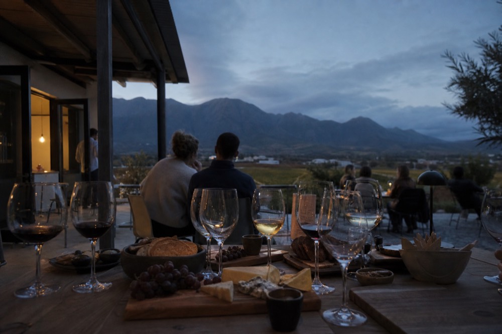 A relaxed group table on a vineyard terrace laid with cheese boards, chocolate and wine glasses, mountains beyond, no readable text, no logos, no recognizable faces