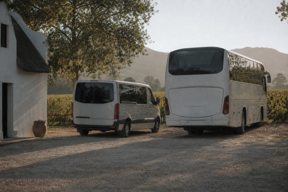 Two touring vehicles, one small van and one larger coach, parked side by side on a gravel estate driveway with vineyard behind, empty, soft daylight, no signage, no text, no people