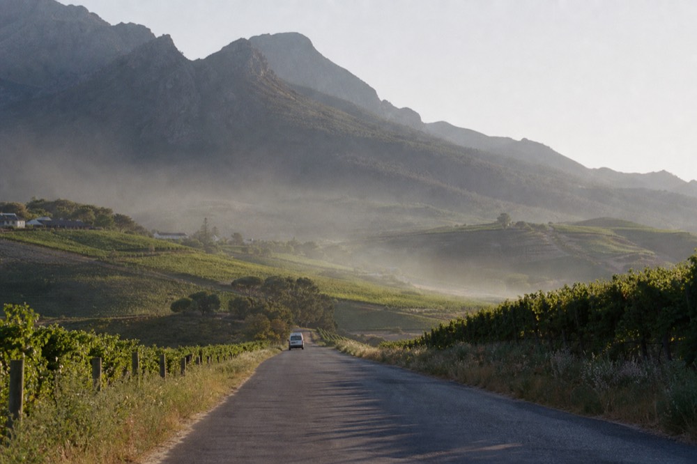 A minibus-scale country road threading between two vineyard valleys with a mountain pass ahead, late afternoon light, no signage or faces