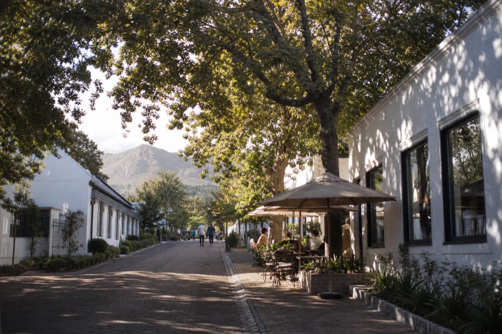 A shaded village side street with gallery windows and cafe tables under oak trees, mountains at the end of the road, no readable signs or faces