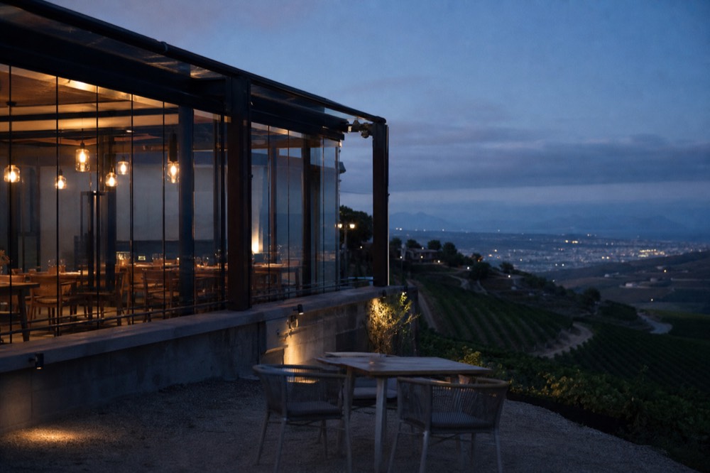 A glass-walled restaurant terrace on a hilltop overlooking terraced vineyards falling away to a distant blue valley and coastline, empty tables, no branding, no text, no people