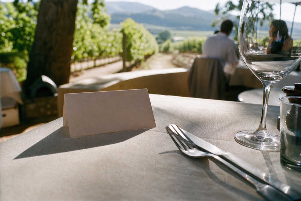 A close-up of a reservation card and cutlery on a linen tablecloth beside a wine glass on a sunlit estate terrace, blurred vines behind, no readable text, no logos, no faces