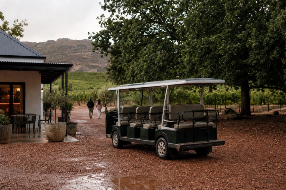 A small open shuttle vehicle parked at a gravel cellar-door forecourt with vineyard and oak trees behind, empty seats, warm afternoon light, no signage, no text, no faces