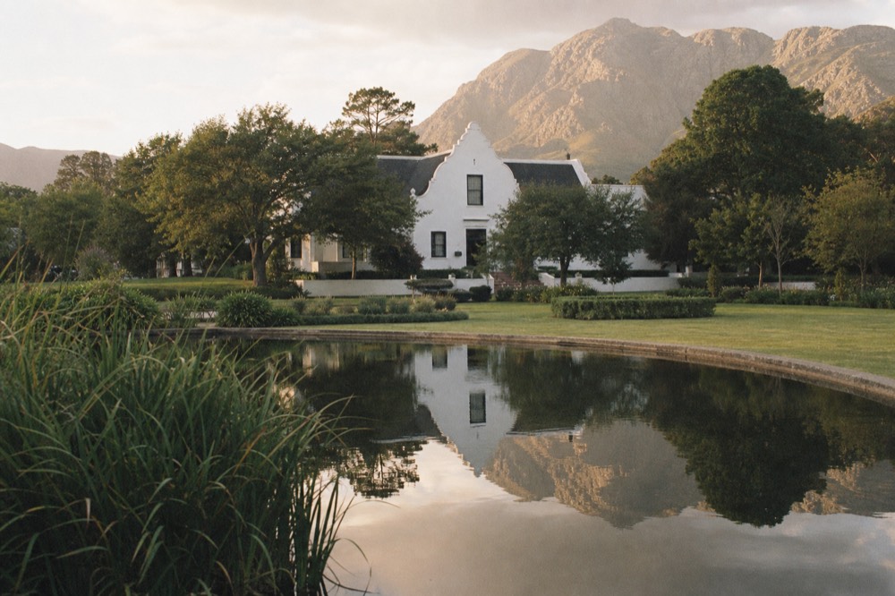 A Cape Dutch estate hotel with white gabled facade reflected in a still garden pond, oak trees and mountains behind at golden hour, no people, no text, no signage