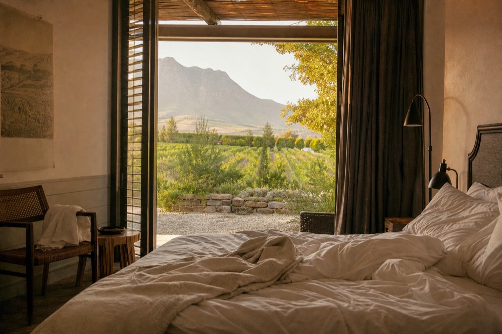 A private lodge suite with an open terrace door framing vineyard rows and mountains, an unmade bed and morning light, no people, no text, no branding