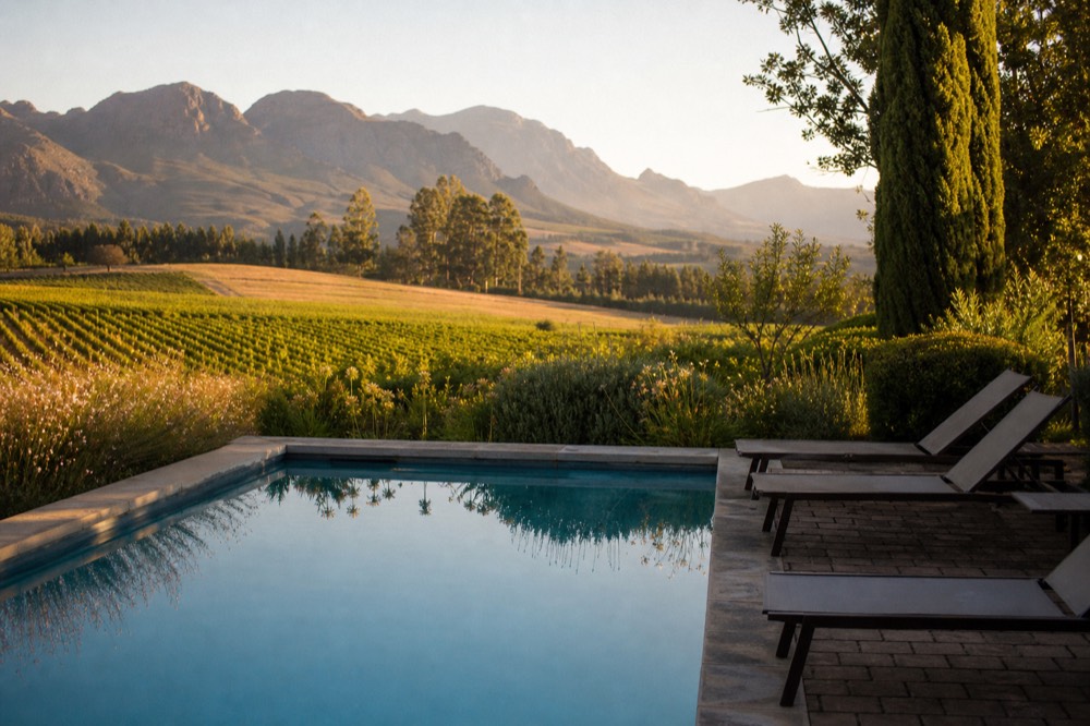 A quiet estate swimming pool and loungers overlooking vineyard rows and mountains on a warm afternoon, no people, no text, no branding