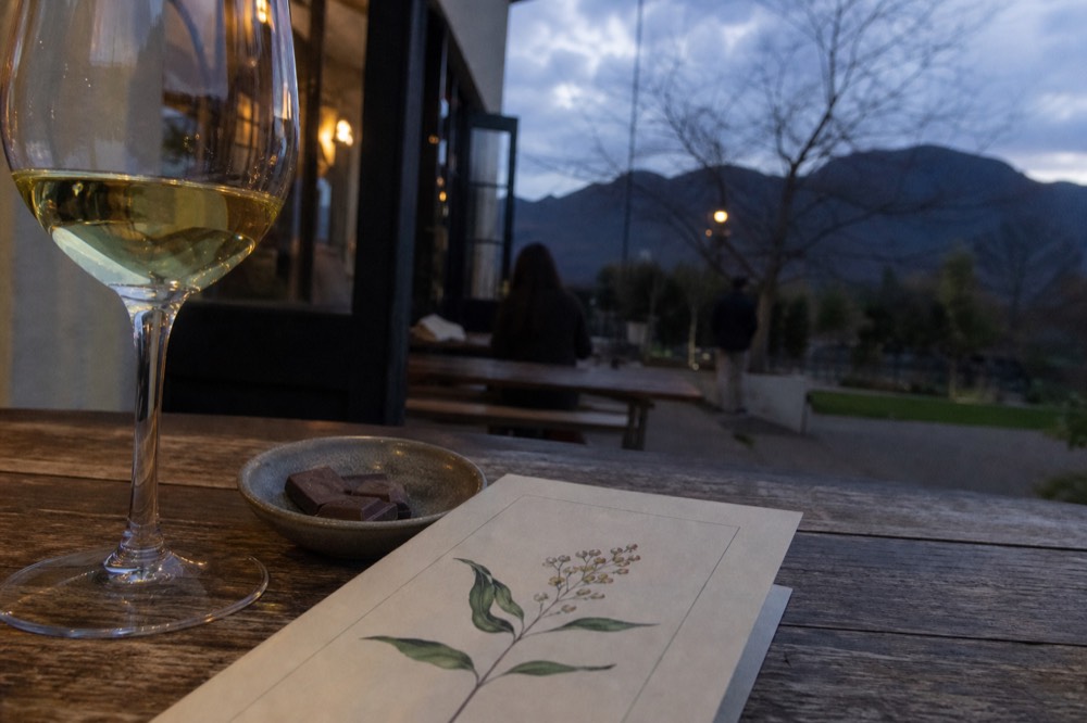 A close-up of a printed tasting menu card on a wooden table beside a glass of white wine and a small dish of chocolate, soft daylight, no readable text, no branding